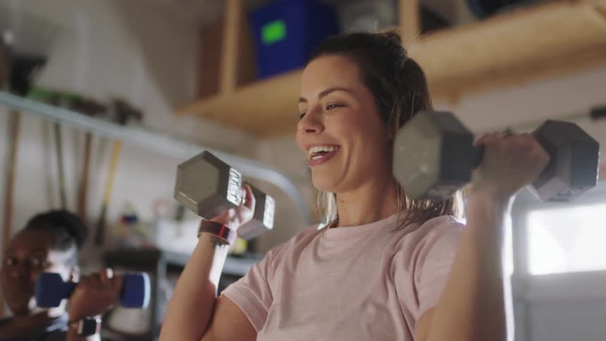 Two women smile at each other while working with hand weights in the garage