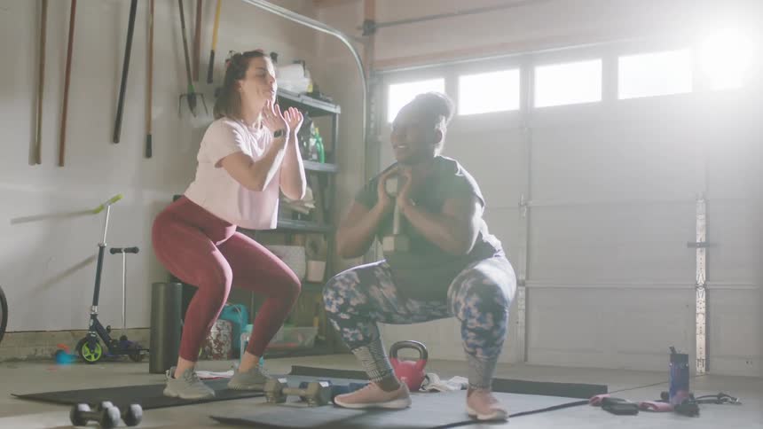 A personal trainer instructs a woman on doing squats with a dumbbell in a garage during the day