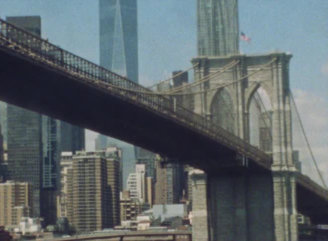 A suspension bridge stretches across the water with an american flag on top and tall city buildings and towers are in the distance