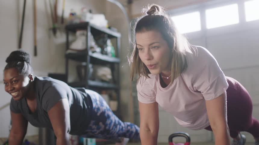 Two women are in a garage smiling as they do push ups on exercise mats