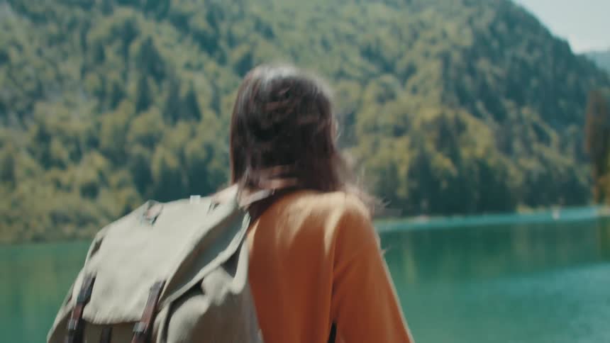 A teenage girl wearing a big backpack is walking along the coast with mountains in the distance as she stops and takes off her backpack
