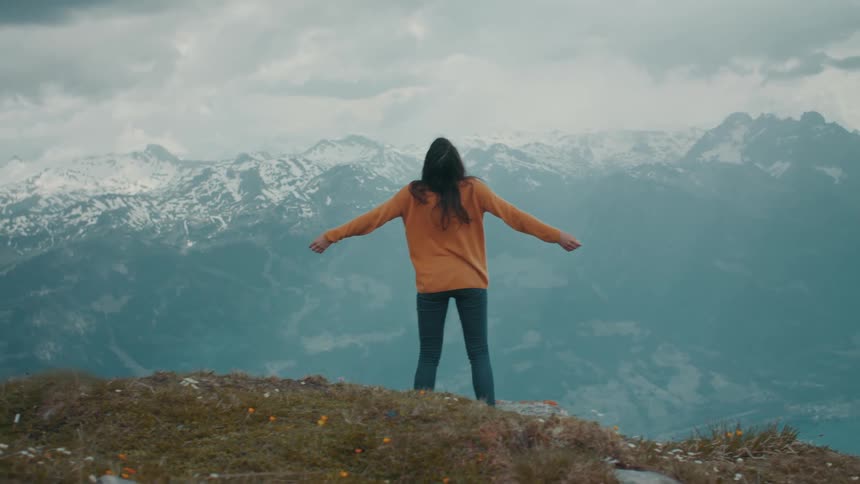 A woman stands in front of a mountain range and stretches her arms to her side as she looks up at the sky