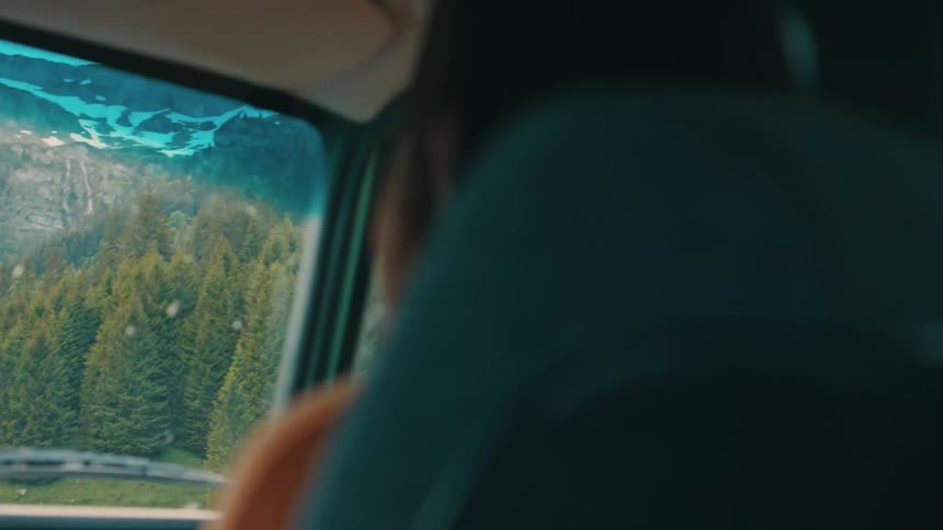 A woman rides as a passenger in a car that is passing a dense forest at the base of a mountain range