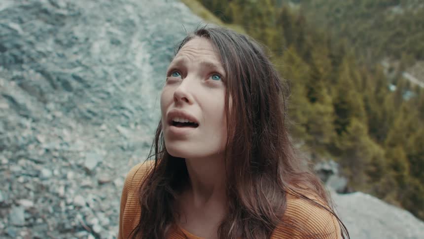 Determined and tired woman with blue eyes brown hair and orange sweater climbs up mountain looking up and breathing