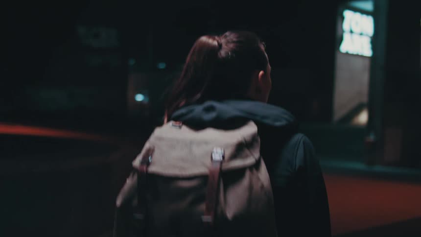 A young woman wearing a backpack walks through a city at night
