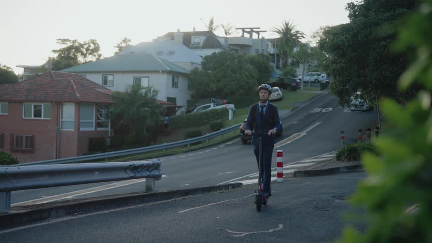A man wearing a helmet rides a scooter on a neighborhood street as traffic moves on the street