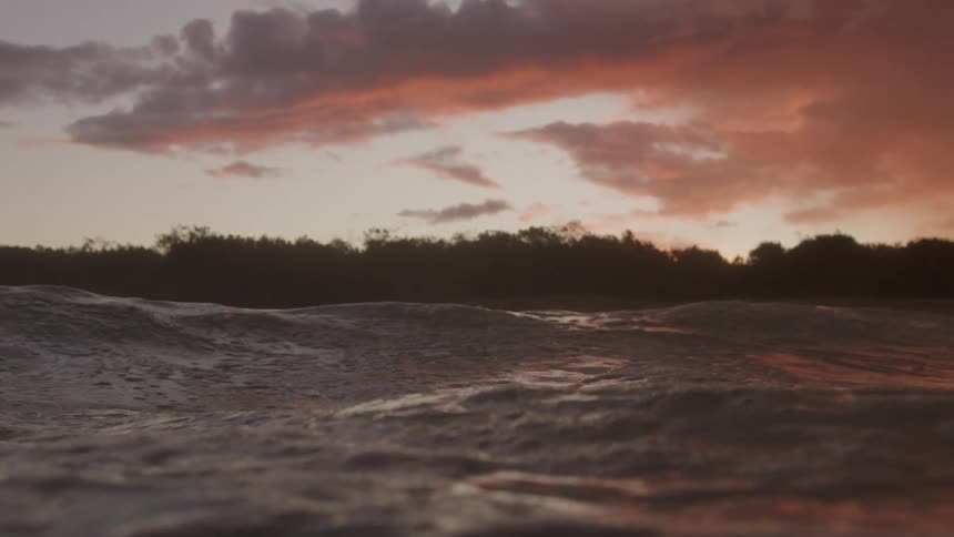 Water waves and ripples as the dark pink clouds above reflect on the surface as it moves overhead
