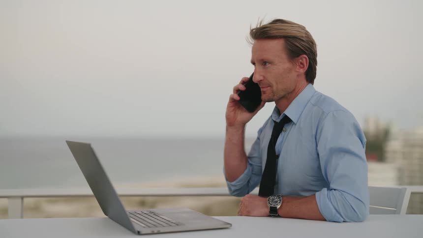 A man answers his phone while working on his laptop outside