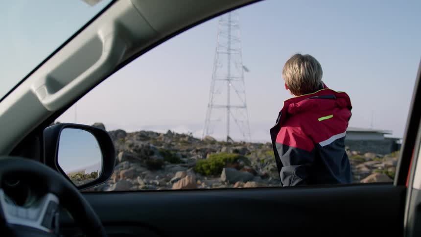 Blonde man wearing black and red jacket talks on smartphone seen from car