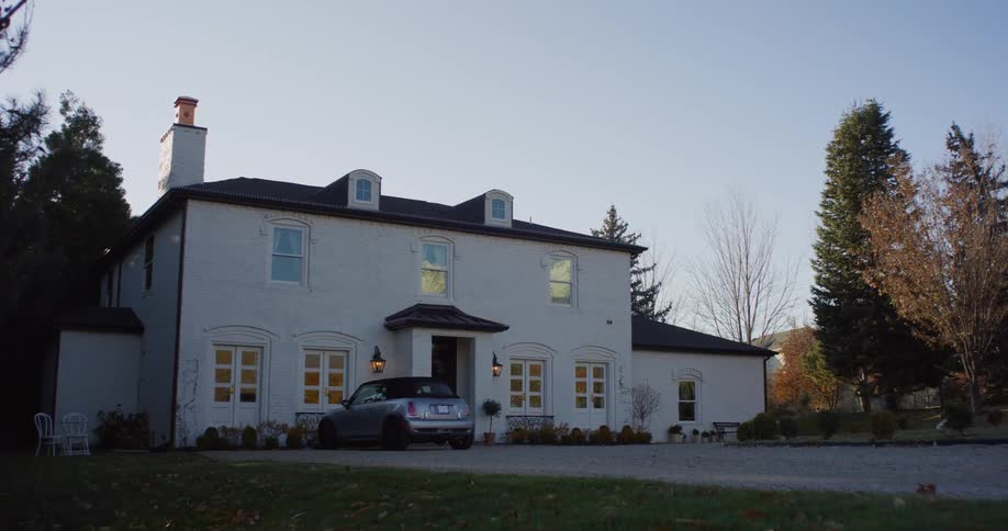 An automobile is parked in front of a large white estate house with trees on the property