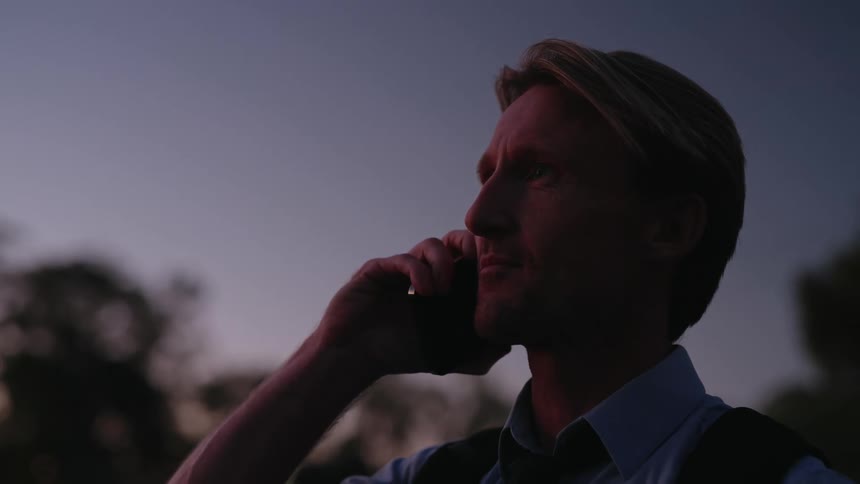 Middle aged blonde man with blue shirt and black vest looks down at smartphone and answers a call outdoors at dusk
