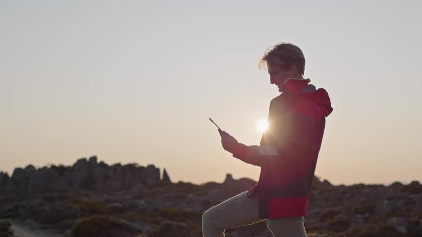 A man is standing outside using a tablet computer as he holds it up the sun shines brightly in the distance