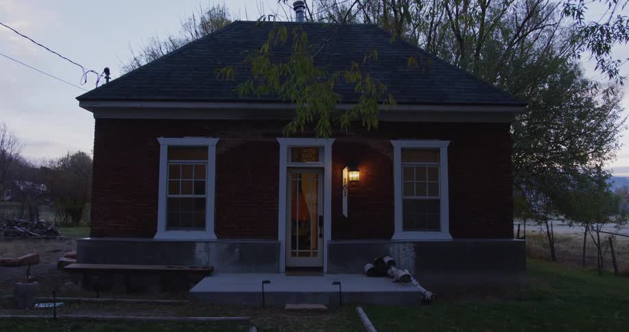 The porch light of a small house is on and another light comes on inside