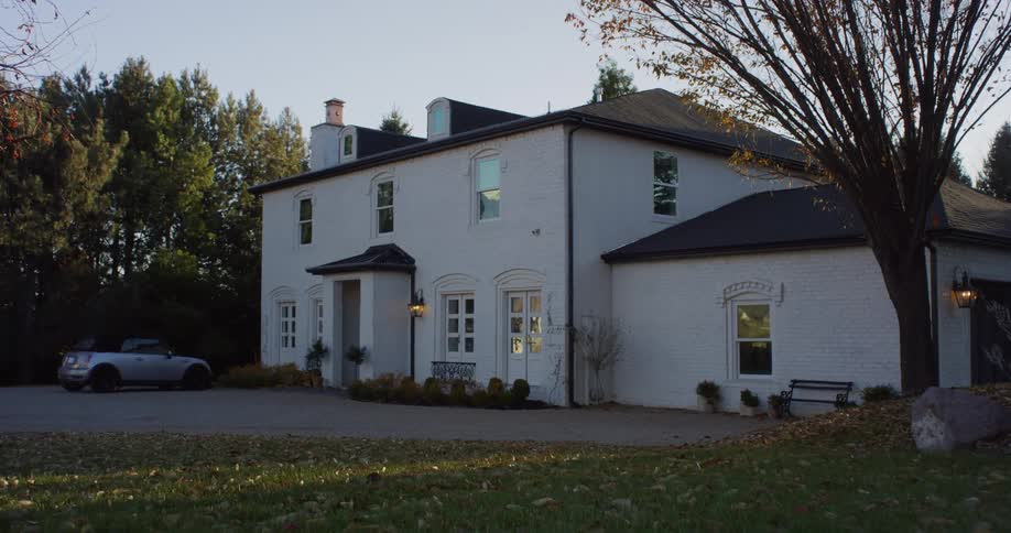 An automobile is parked outside a large house as the sun shines on the trees under a clear sky