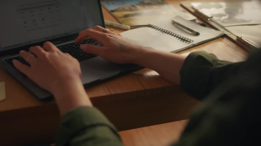 Woman sits at desk making notes and typing on a laptop