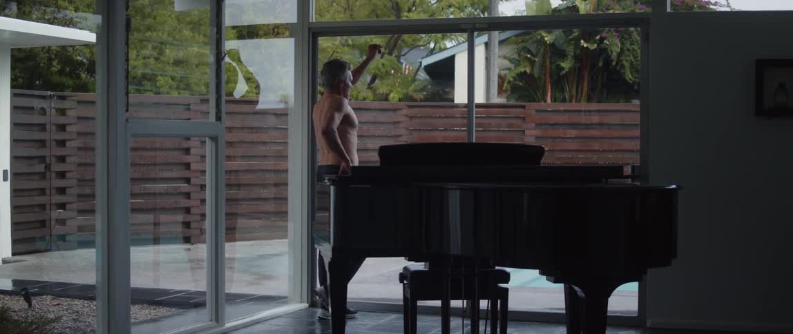 A man walks past a piano in his house to look out the window