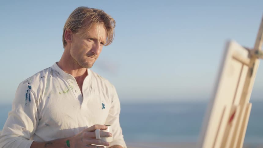 A painter drinks form a mug while looking at his painting