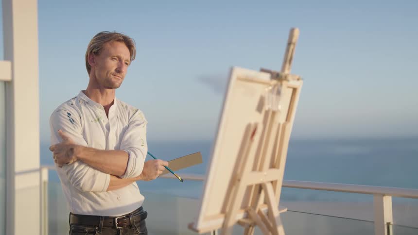 A man stands on a balcony on the shore and paints on a canvas