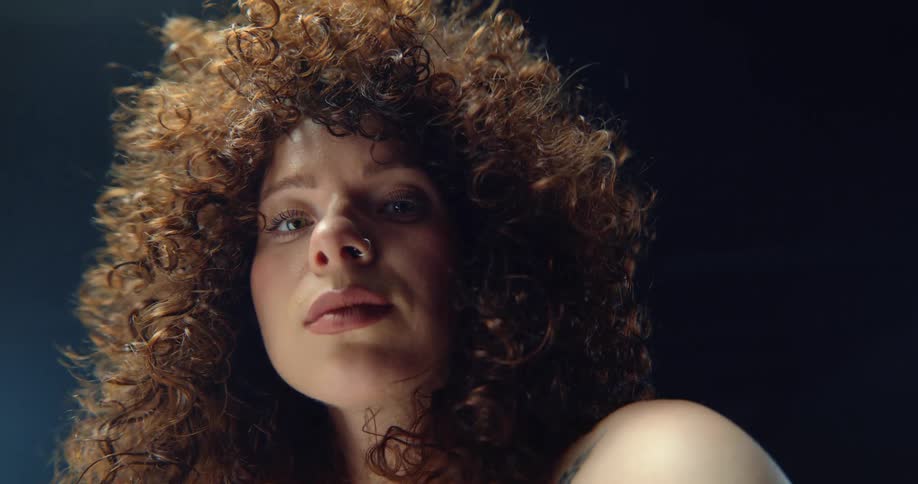 Female model with curly hair piercing and tattoo looks down at camera in the studio
