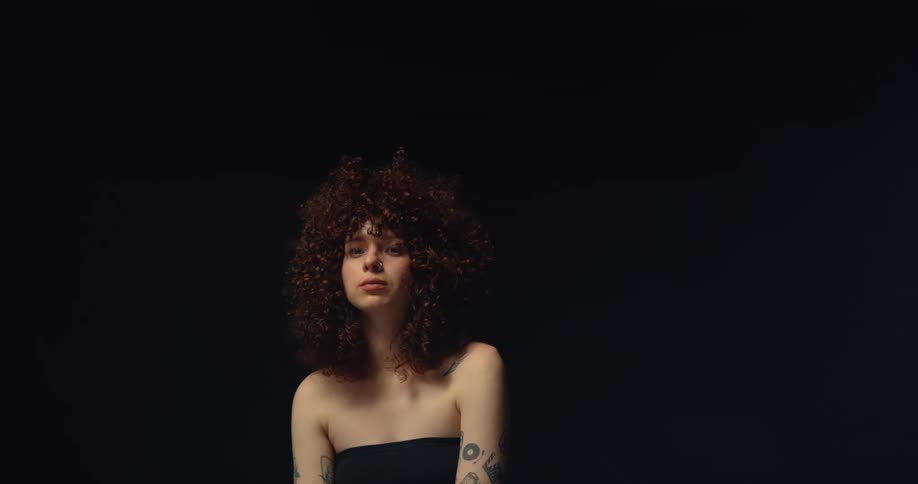 A woman with curly hair is sitting and posing in front of a black background