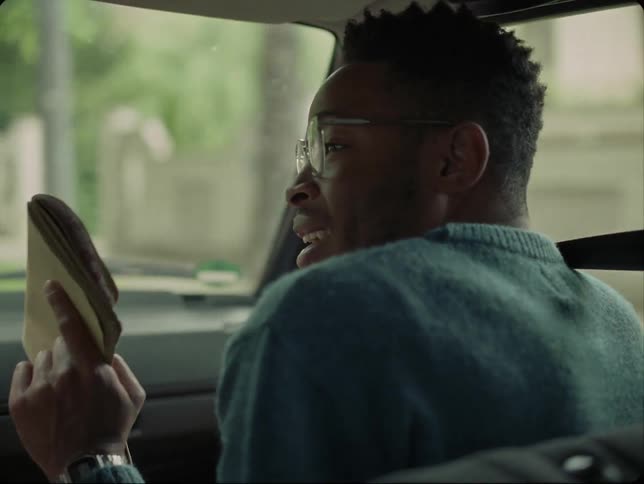 Man with glasses and blue sweater argues with person showing notebook in moving car on the street