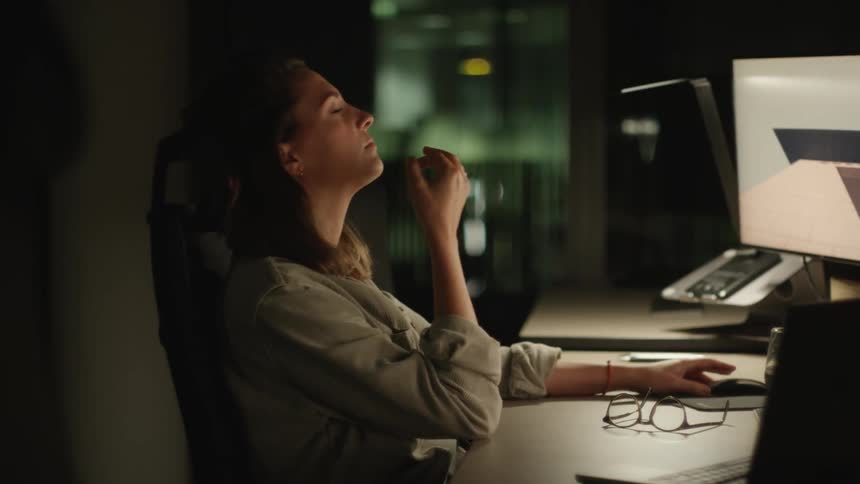 A woman sits at her desk in her office at night and rubs her eyes and leans back in her chair