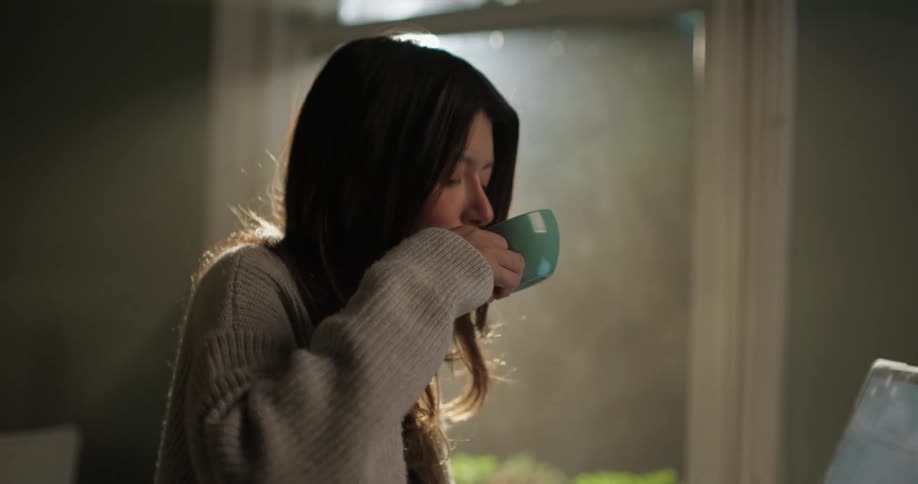 Woman takes short break to sip drink while typing on laptop near cafe window