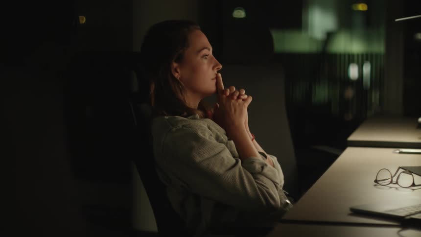 A woman sits at her office desk at night and rubs her neck as she looks at her computer and thinks