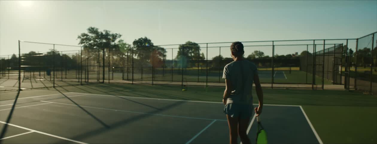 Female tennis player takes place on one side of tennis court as traffic passes on road at edge of park