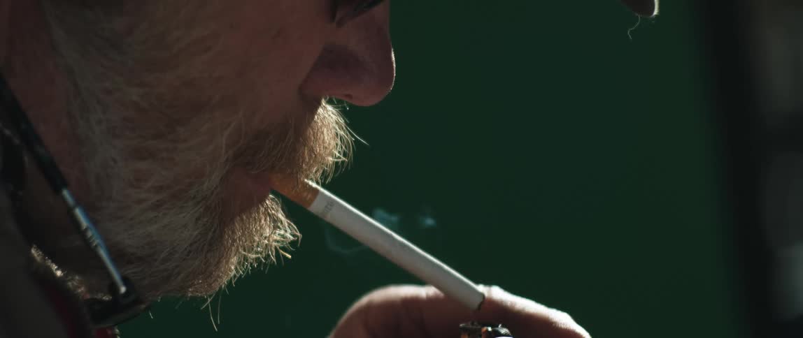A bearded man uses a lighter to light a cigarette that hes holding in his mouth