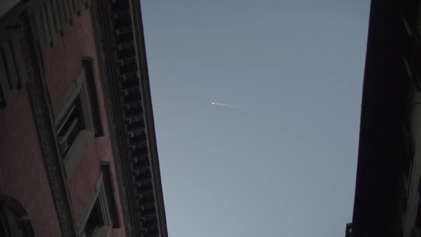 A plane flies high above two buildings