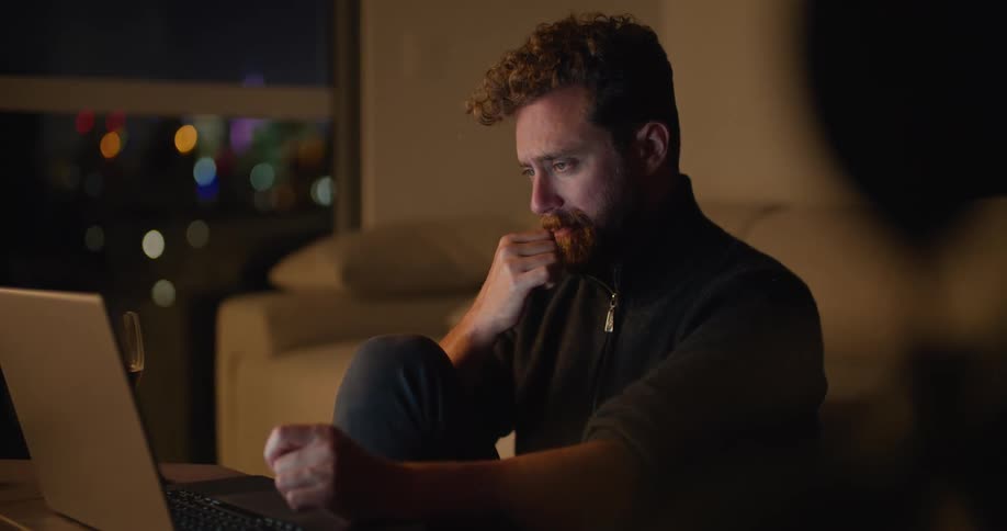 A man thinks for a moment before beginning to type on a laptop while sitting on the floor at a table indoors at night