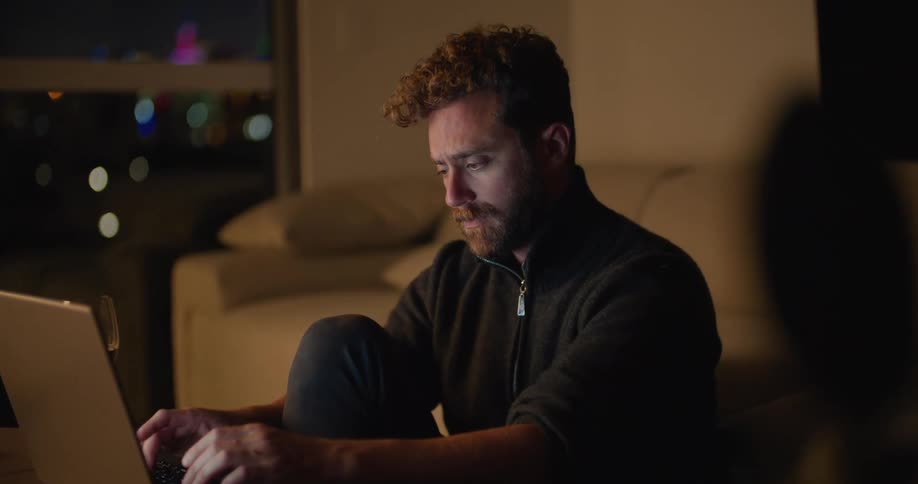 A man types on a laptop while sitting on the floor before a coffee table indoors at night