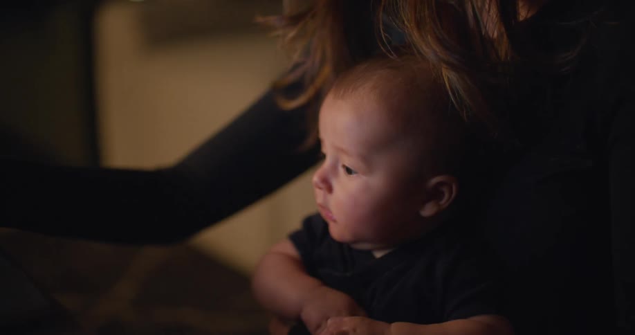 Woman holding a baby uses a laptop in the living room at night