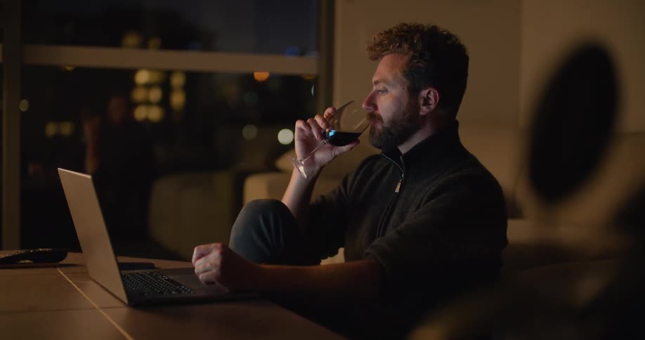 A young white man is sitting down and using his laptop while drinking some wine