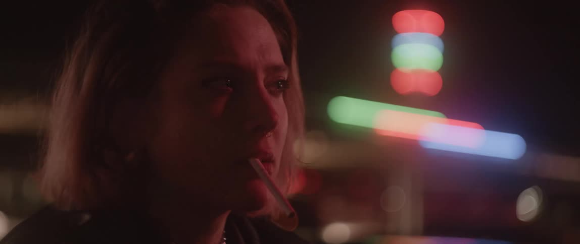 Blonde woman smokes while looking around at night as nearby neon lights flash