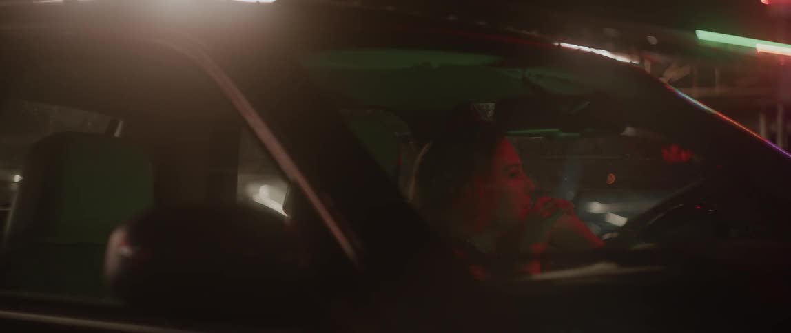 Woman sitting on a car parked near a store at night drinking soda