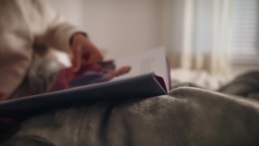 Woman reads a book while resting on the bed under a thick blanket at home
