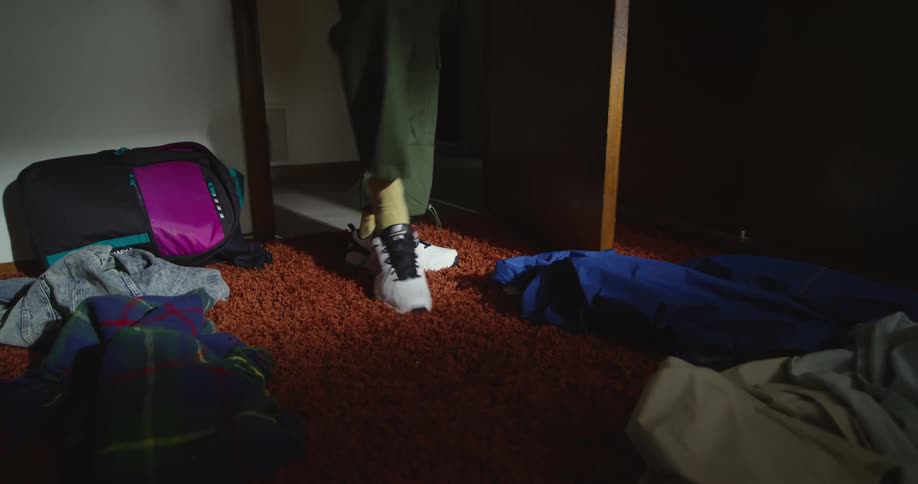 Person kicks off their sneaker as soon as they enter room with blankets and clothes strewn across carpeting
