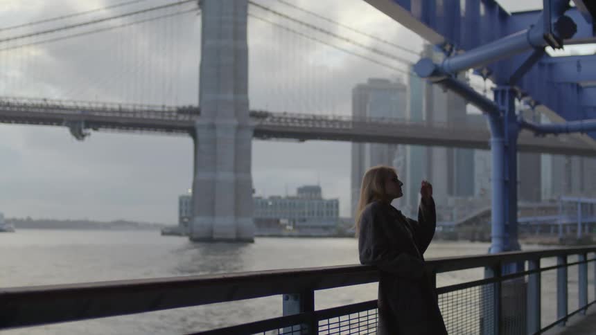Woman leans against rail on waterfront across water from suspension bridge and downtown skyscrapers