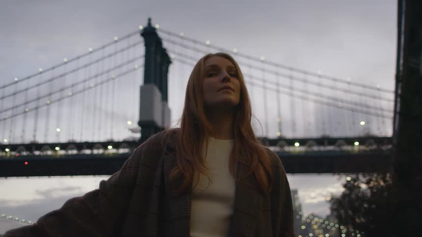 Long haired blonde woman standing in front of a suspension bridge in the city looks around