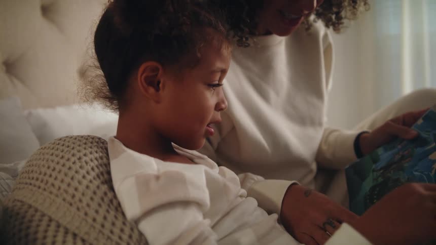 A black child and black mother reading from a book on a bed