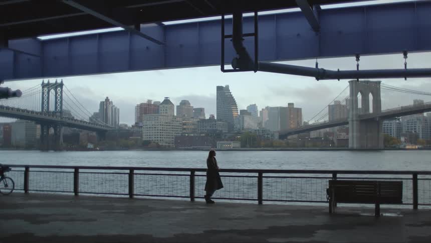 Woman stands near rail across water from downtown skyscrapers