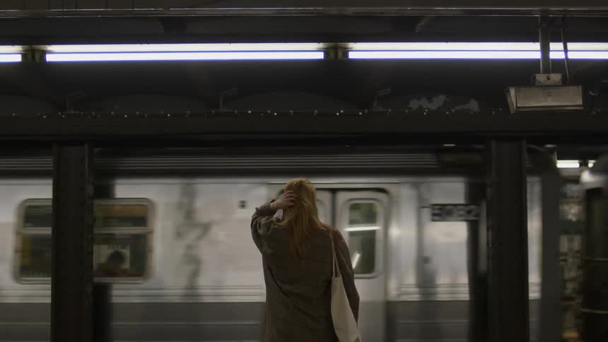 A woman wearing a trench coat with her purse over her shoulder is standing in the subway watching a train go by