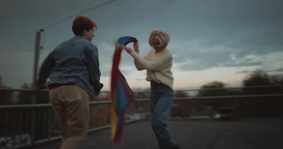 Two women play chase as one carries a rainbow banner with city in the background