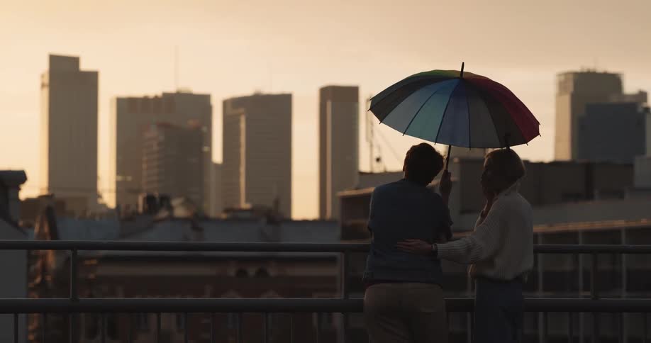 Two people stand close together under rainbow colored umbrella looking at city skyline under a red sky