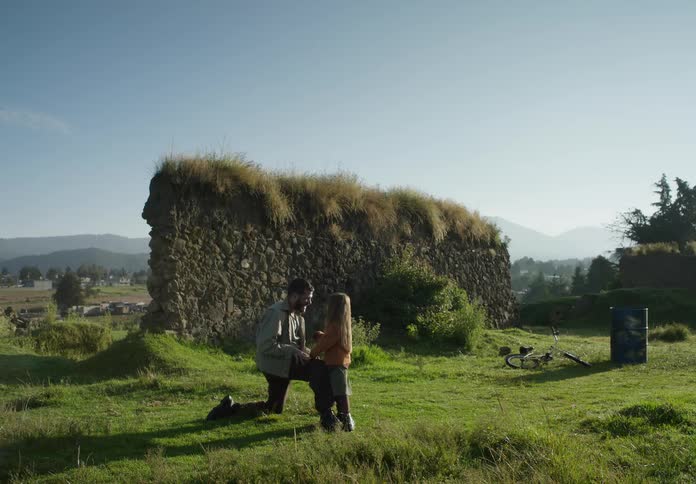 A man is outside on one knee in the grass with his young daughter near ruins of a wall and there are mountains in the distance