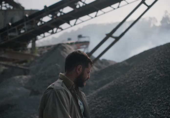 A male worker stands outside at a quarry near some machinery as he decides what to do