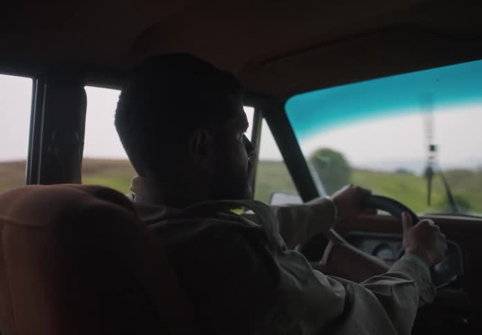 A man driving an automobile holds the steering wheel tightly with both hands as he travels on a rural road and the windshield wipers move across the glass