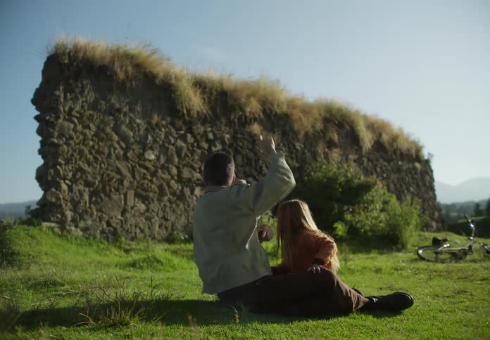 A man is sitting outside in the grass with his young daughter near ruins of a wall as he lights a firecracker and throws it then he gives her a hug